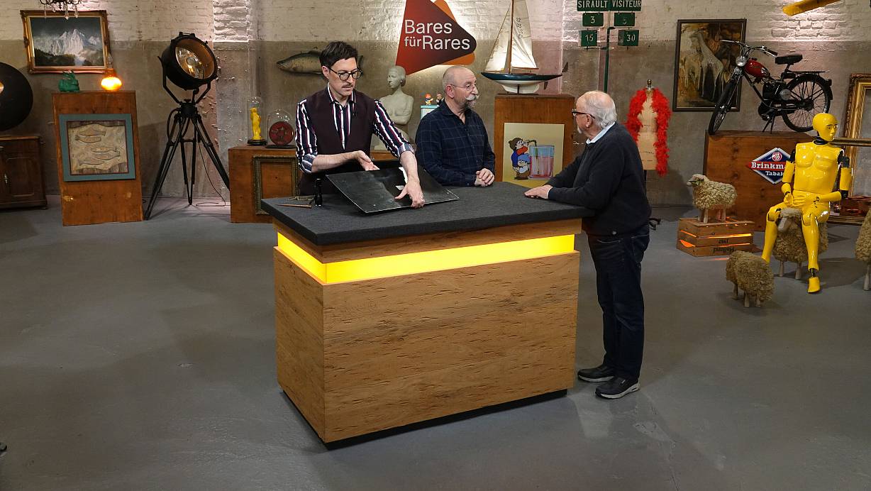 Experte Colmar Schulte-Goltz (v.l.) begutachtet bei "Bares für Rares" das Miele-Emailleschild, während Moderator Horst Lichter mit dem Harsewinkeler Georg Terlutter redet. | © ZDF/Warner