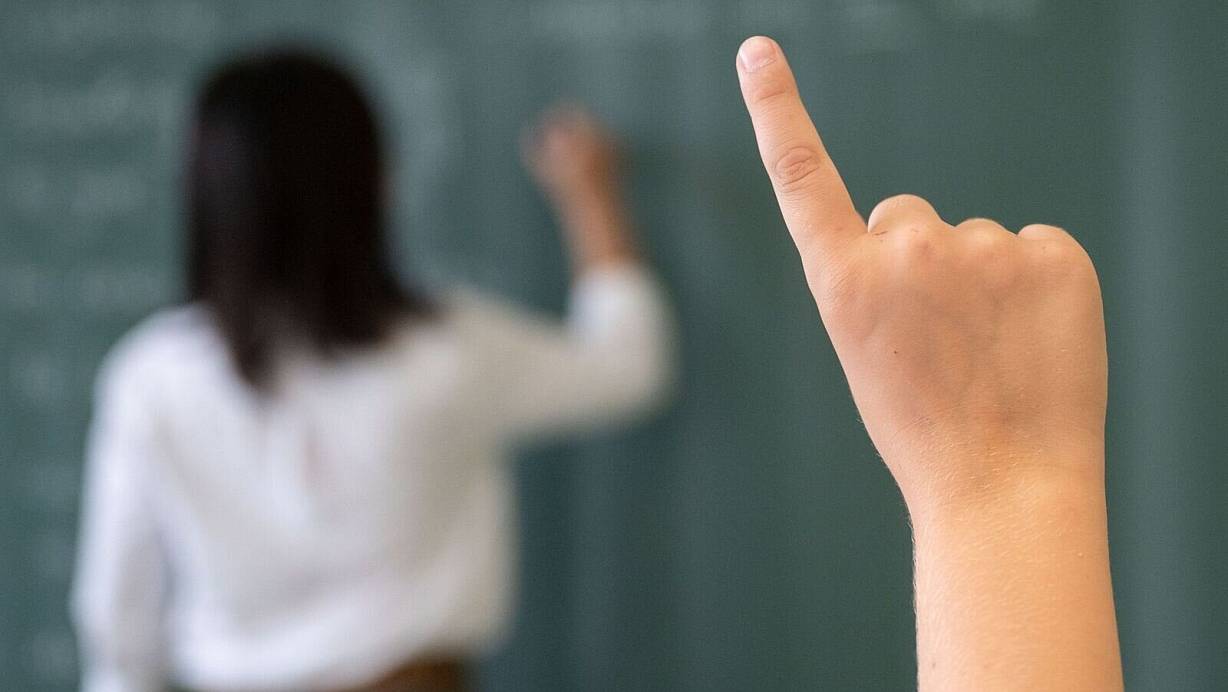 Ein Schüler meldet sich, während die Lehrerin an die Tafel schreibt. | © Marijan Murat/dpa/Symbolbild