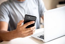 Mann hält vor einem Laptop sein Smartphone in der Hand - Zacharie Scheurer/dpa-tmn