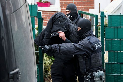 Polizeibeamte führen einen Tatverdächtigen aus einem durchsuchten Haus. - © Christoph Reichwein/dpa