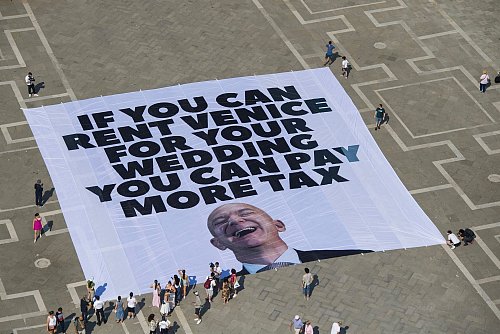 Das Transparent zeigt ein Foto des Amazon-Gründers, auf dem steht: «If you can rent Venice for your wedding you can pay more tax». - © Greenpeace/Greenpeace/AP/dpa