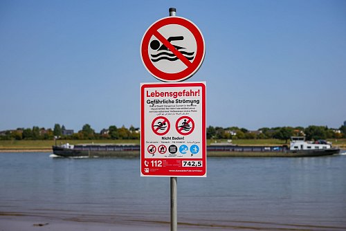 Schilder, die auf die Gefahren beim Schwimmen im Rhein und auf das ausgesprochene Badeverbot hinweisen, an einem Schildermast am Rheinufer.(Archivbild) - © Christoph Reichwein/dpa