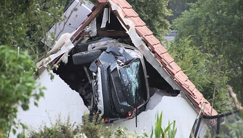 Das Auto liegt nach einem kurzen Flug seitlich in der Mauer einer Scheune. - © -/Moonshine Media/dpa